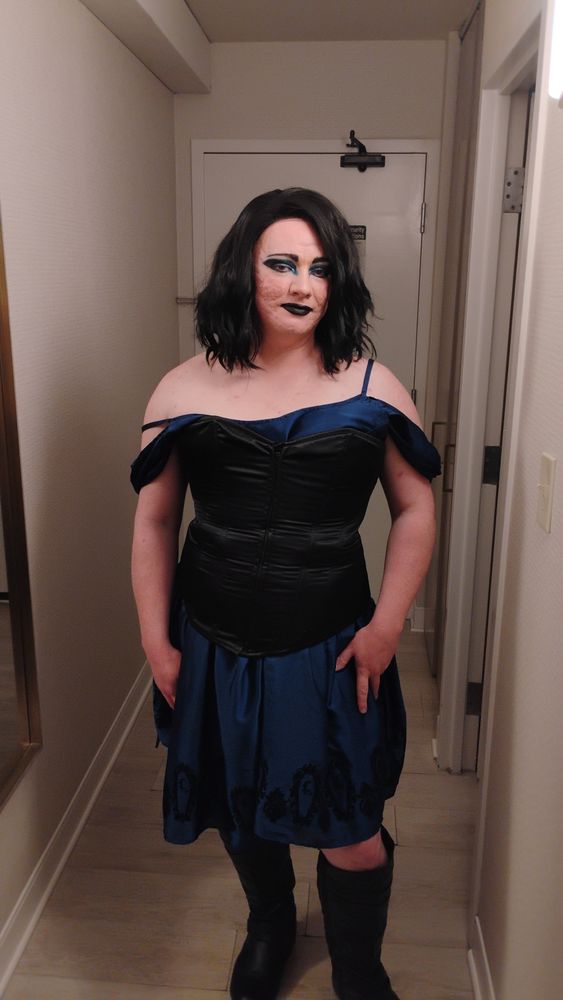 Drag queen standing in a hotel hall wearing: black wig, a black corset over a blue dress with black embroidery, and black knee-high boots. She has black lipstick and blue eyeshadow.