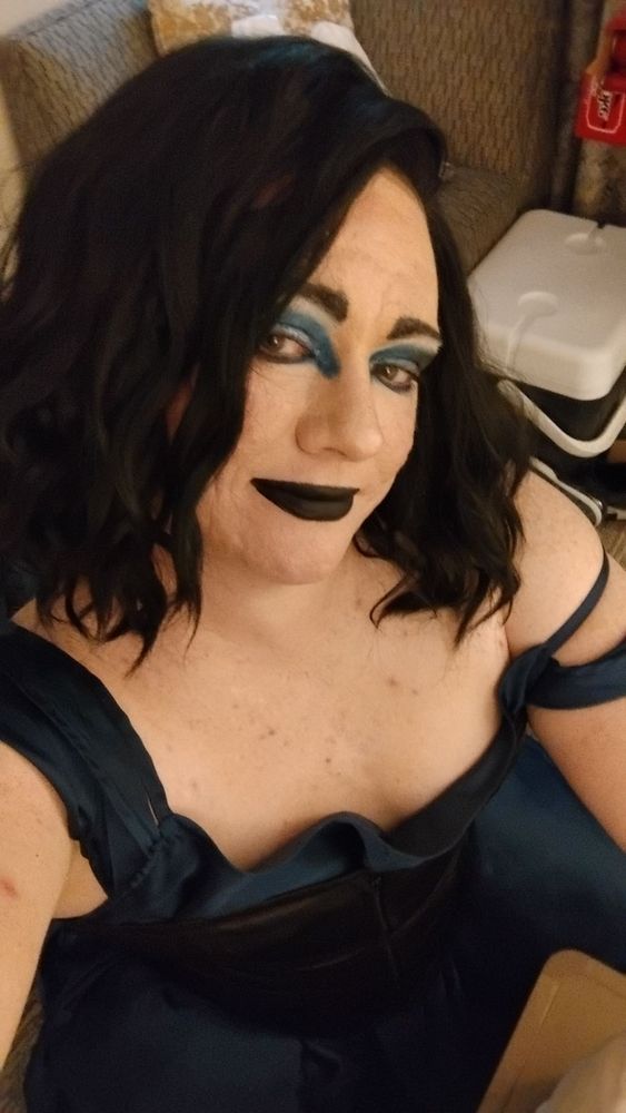 Shot from above selfie of a drag queen in: black corset over a blue dress, a black wig, and blue eyeshadow with black lipstick 