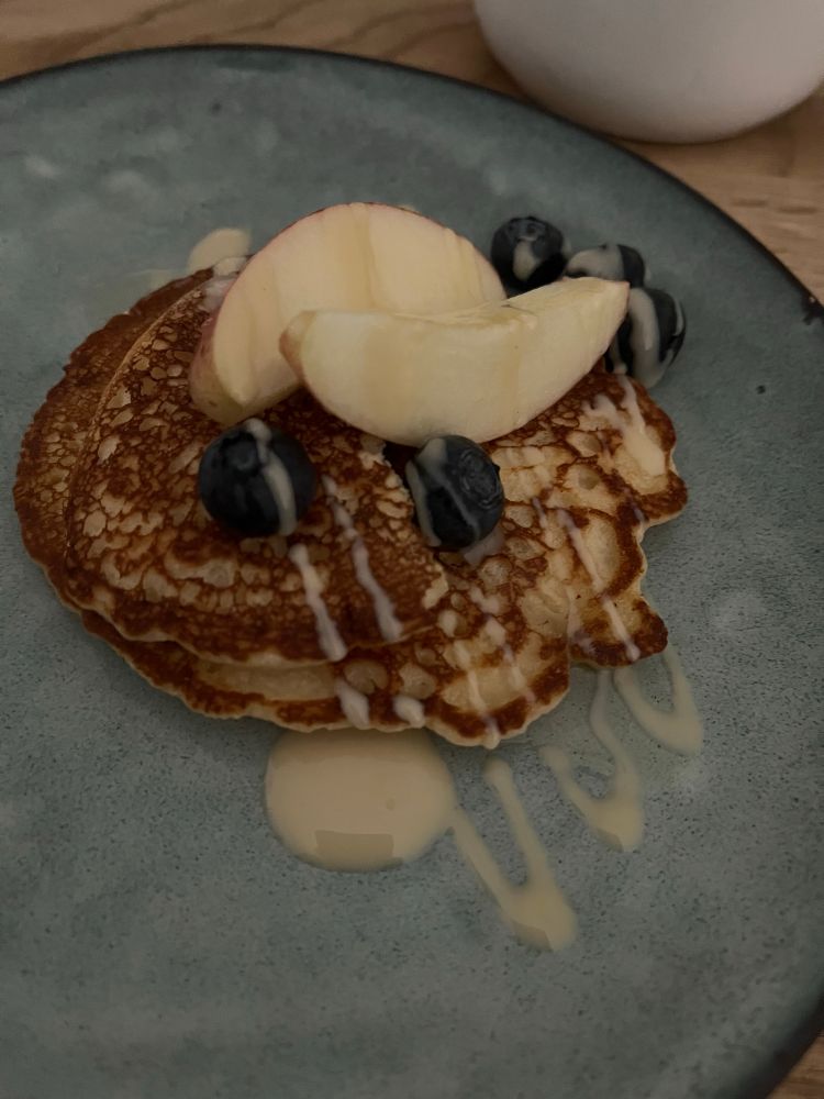 Nahaufnahme eines blauen Desserttellers. Darauf liegen zwei Pancakes, Apfelscheiben und Heidelbeeren mit Mandelmus