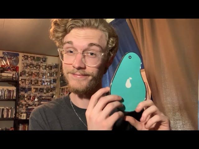 A man holding up a pear-shaped phone, with the backside of the phone facing the camera. The back of the pear-phone is teal-colored. There is a white pear logo in the center of the backside of the phone, meant to resemble the Apple logo. On the top edge of the pear-phone, where the smaller side of the pear-phone is, there is one single camera lense visible on the top.