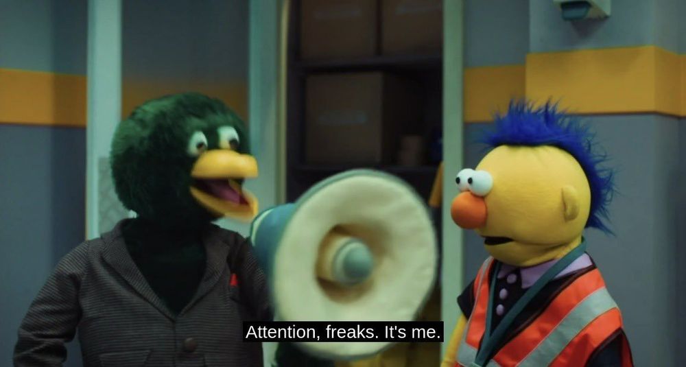 A green duck puppet speaks into a megaphone next to a blue-haired puppet in a safety vest. Caption reads: “Attention, freaks. It’s me.”
