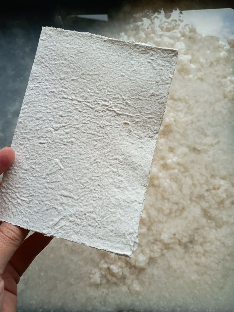 Handgeschept wit papier met op de achtergrond de papierpulp in een bak met water 