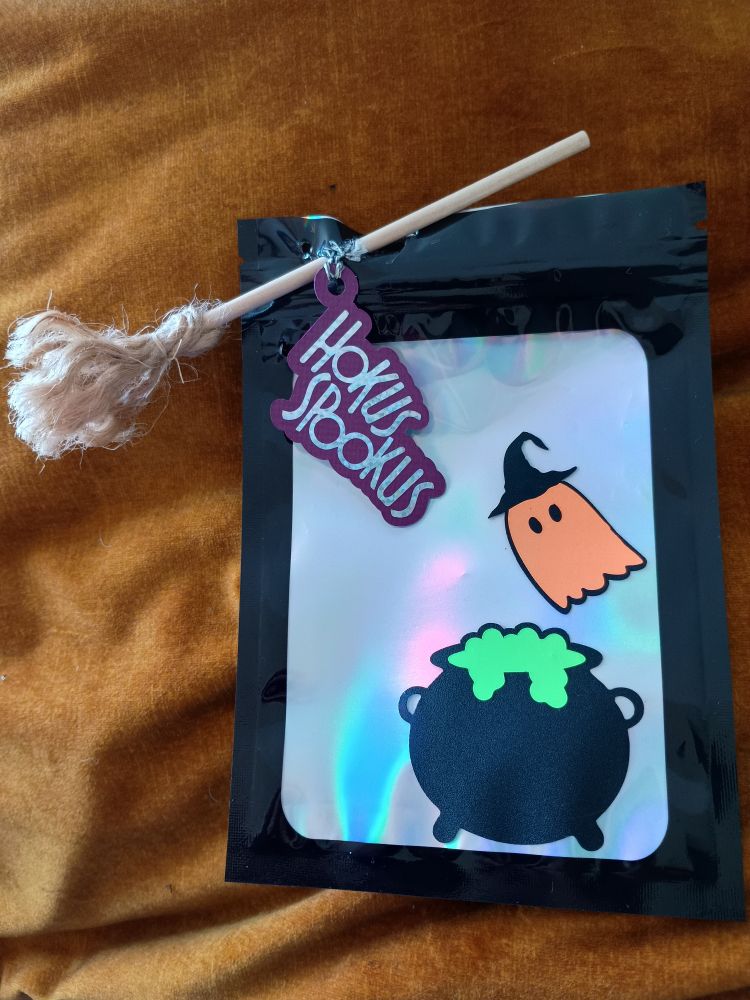 Zwart snoepzakje met doorzichtig venster en iriserende achtergrond. Versierd met stickers: heksenketel en groene smurrie en oranje spook met heksenhoed. In de rechterbovenhoek van het zakje zit een een bezem door het zakje. Bezem is gemaakt met een houten stokje en touw en er hangt een papieren tag aan met de tekst "hokus spookus"