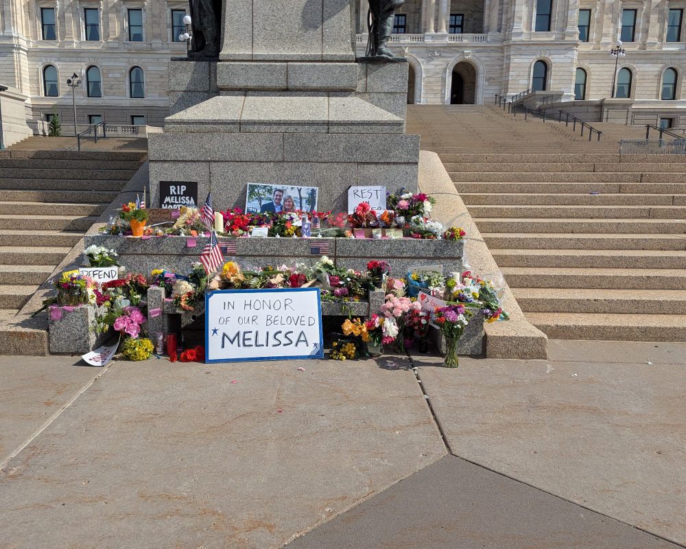Memorial with flowers at the base of the state Capitol for Rep. Melissa Hortman. Sign reads "in honor of our beloved Melissa"