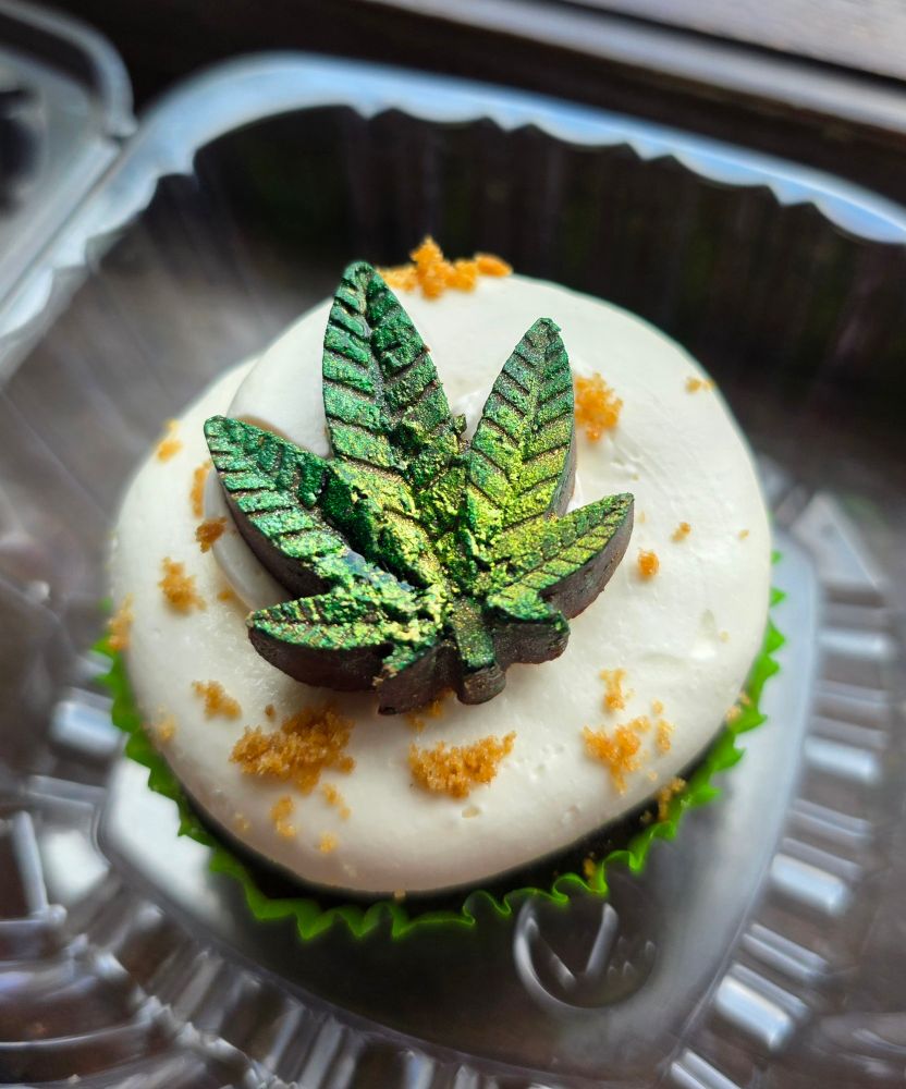 A smore cup cake with a cool shiny pot leaf candy topping, white marshmallow frosting, and a sprinkling of brown sugar to look like kief or powdered blonde hash