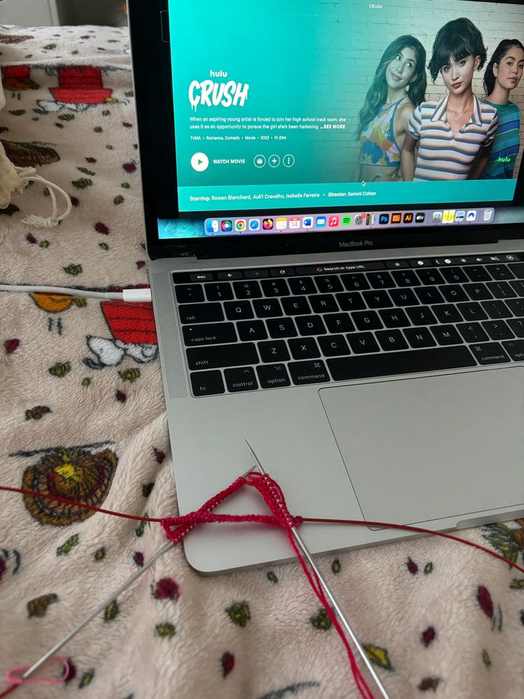 red yarn cast on to a long fixed circular knitting needle, leaning against a laptop with hulu open