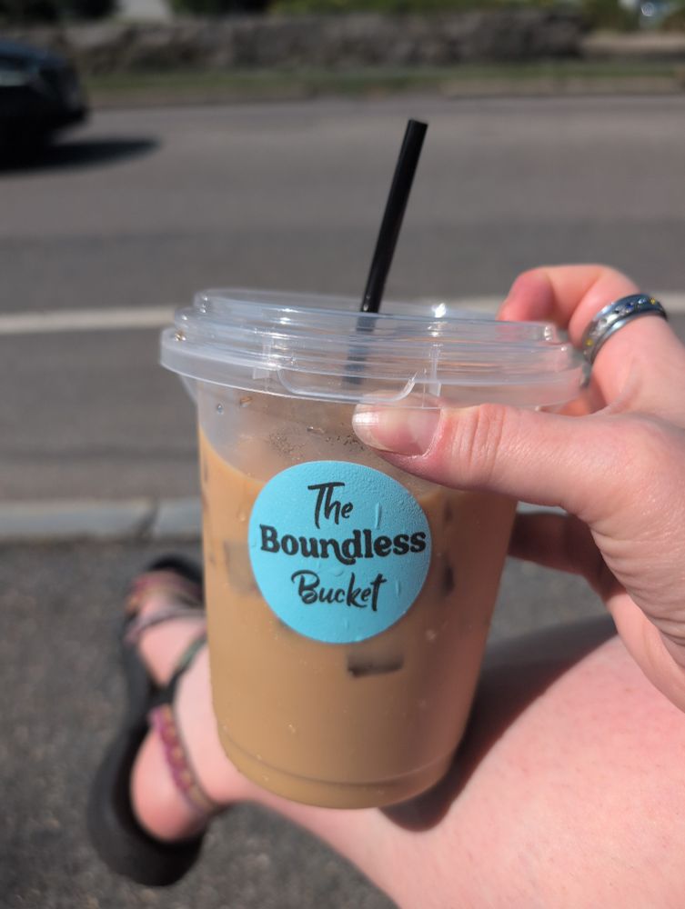Hand holding a very large iced coffee with the label "The Boundless Bucket"