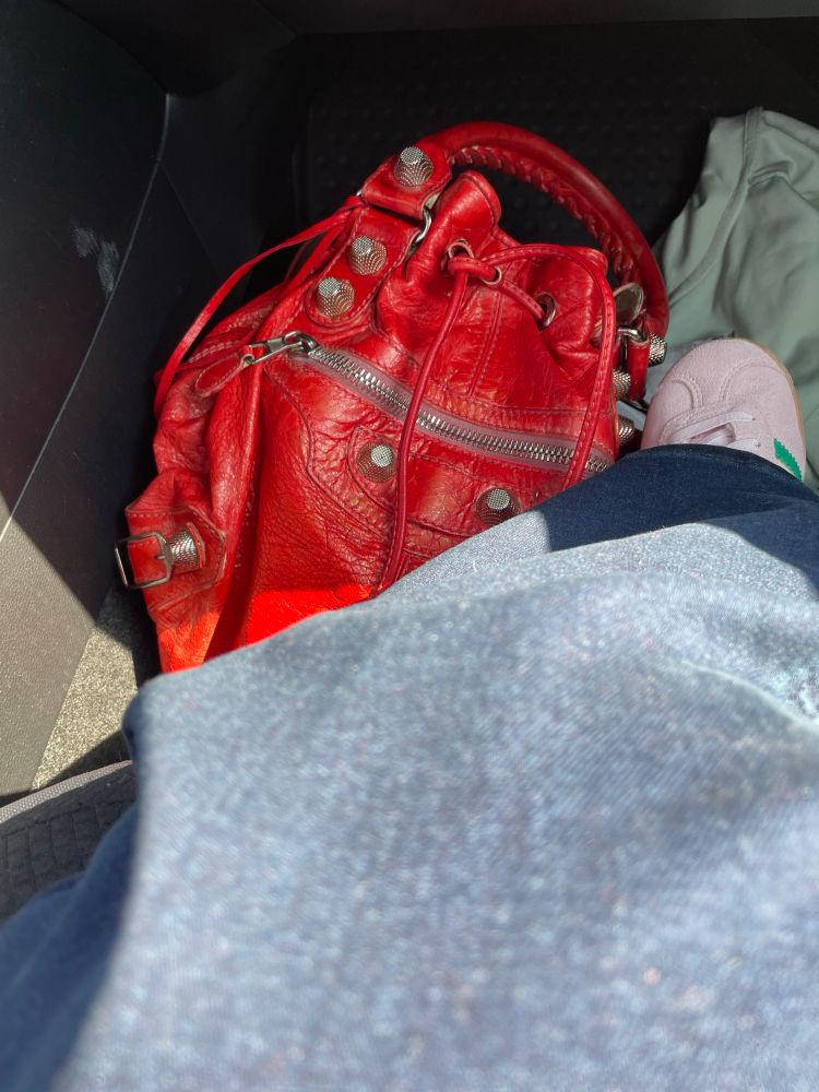 Red leather handbag with metal studs and zippers rests on a car floor mat. Nearby, a pink shoe and some denim