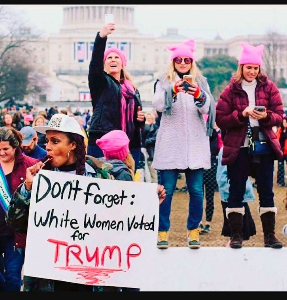 On the right, three white women in “pink pussy” hats stand on a platform and use their phones.

On the left, a Black woman passes by them with a sign that reads: “Don’t forget: White Women Voted for TRUMP”