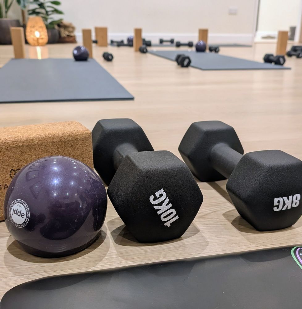 A selection of weights, yoga bricks and pilates balls sit on a pale wooden floor in a white room with plants, lamps and yoga mats. 