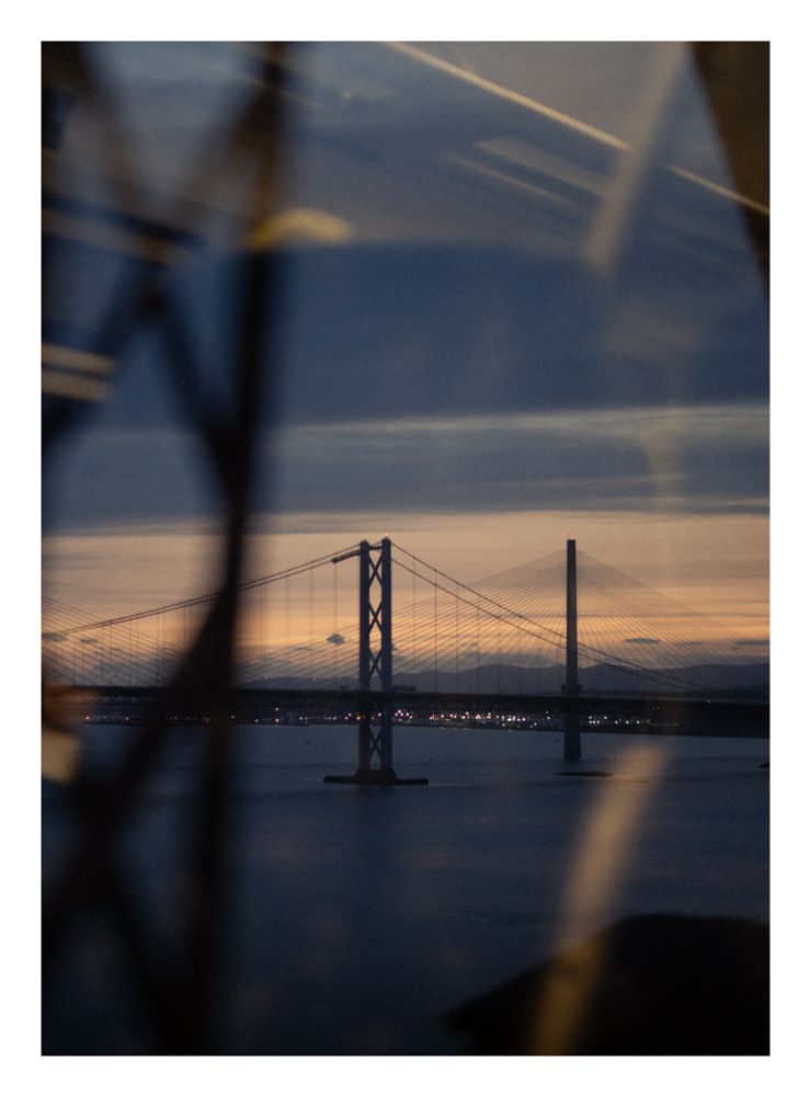 A photograph captured at dusk from the window of a moving train.
In the dark blue evening, a bridge is visible in the distance against the backdrop of a post-sunset sky. Reflections from the train's interior can be seen in the foreground, along with a blurry and fast-moving piece of the architecture on which the train is rolling.