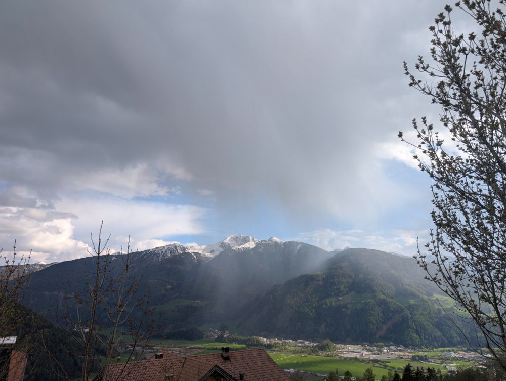 Man sieht die Fallstreifen des Niederschlages von einem Schauer über einem am Gipfel schneebedeckten Berg auf der anderen Seite eines Alpentales.