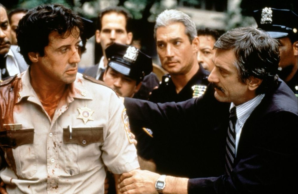 Movie still photo from the movie “Copland.” Sylvester Stallone dressed in a cop uniform with blood on his shirt looking at Robert Deniro who is leaning towards him