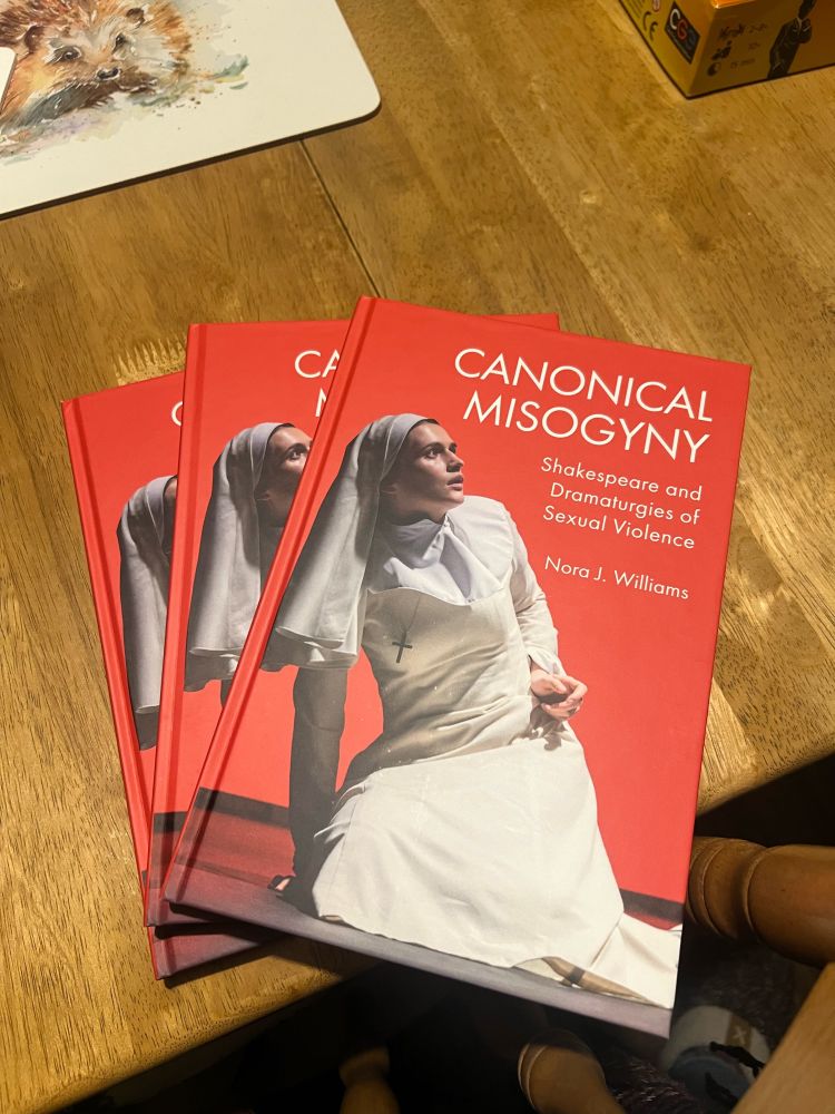 Author copies fanned out on a wooden dining table. Book title is Canonical Misogyny: Shakespeare and Dramaturgies of Sexual Violence by Nora J. Williams. It has a vibrant red background, with an image of an actor playing Isabella in Measure for Measure