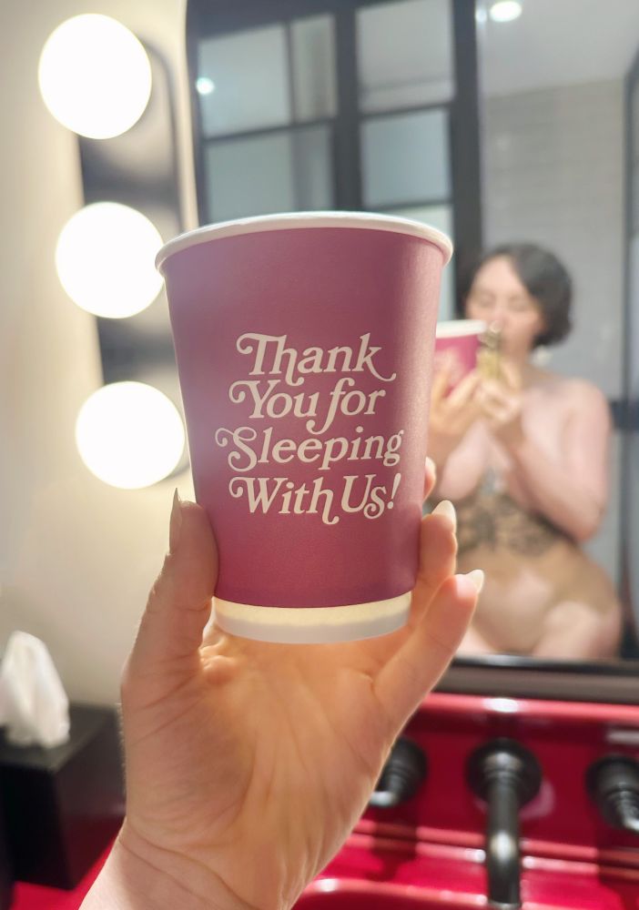 Rose holds a coffee cup in her well-manicured hand that is pink and says, "Thank you for sleeping with us!" in a jaunty serif font. In the background there's a mirror, where you can see a reflection of Rose in her finest. She is pale and curvy with a black ink tattoo on her ribcage of roses, and has a chin-length brunette bob.