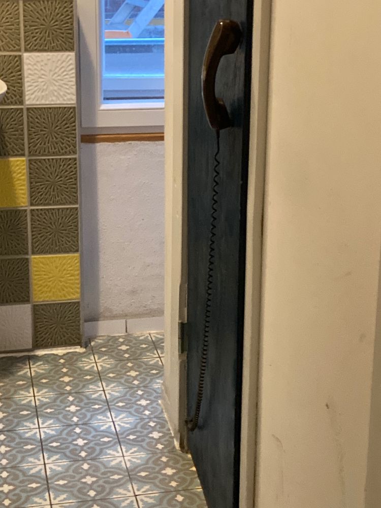 Eine Tür in einer öffentlichen Toilette. Als Klinke ist einen Hörer von einem Schnurtelefon angebracht.