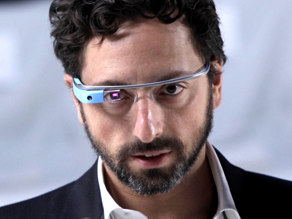 A man looking at the camera wearing a pair of really dorky looking Google Glasses with a camera built in to the side and a lense Infront of the wearers right eye glowing.