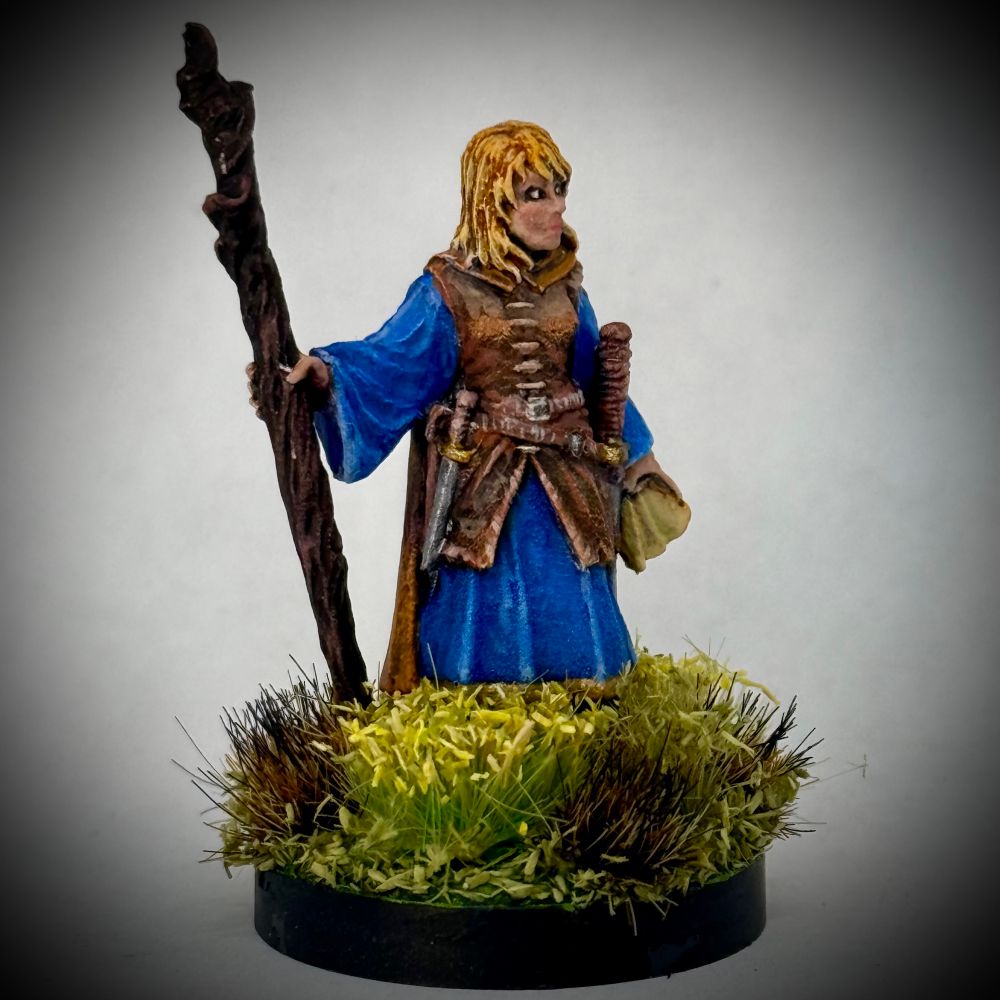 A painted miniature of a female cleric holding a wooden staff and wearing a leather tunic over blue robes. She has pale skin, blond hair, and carries a scroll in her left hand. The base is covered in textured green grass tufts with a patch of scorched grass near her feet. 
