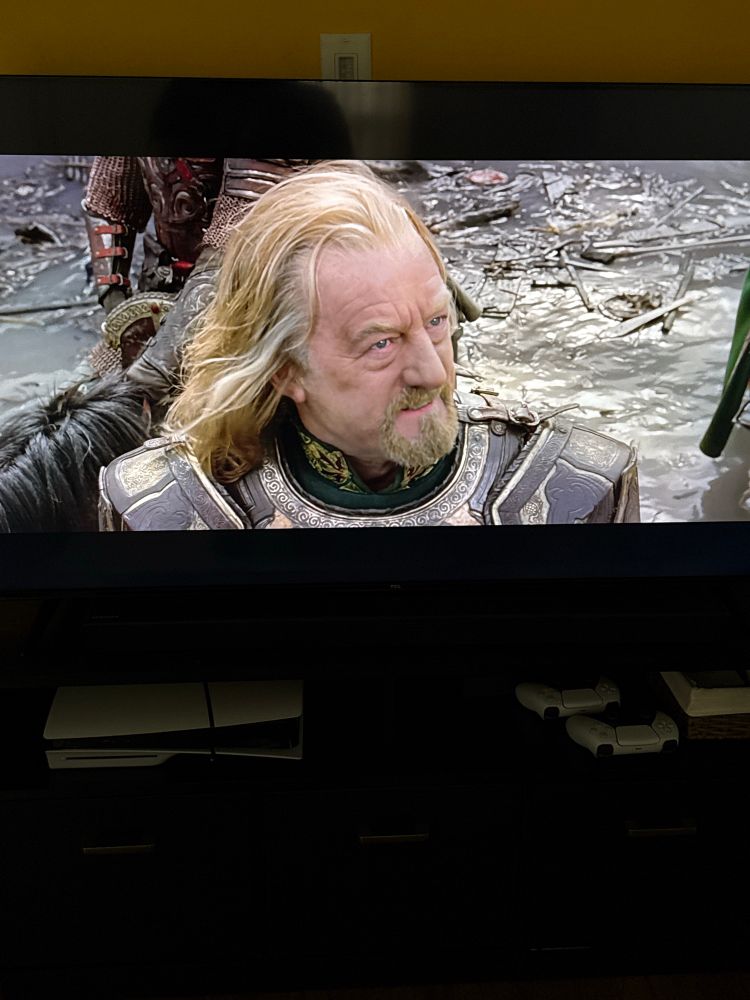 A tv showing a scene from the movie The Lord of the Rings: The Return of the King where King Théoden talks to Saruman at Orthanc. The TV sits on a black entertainment center, which has a PlayStation 5 and two white controllers. 