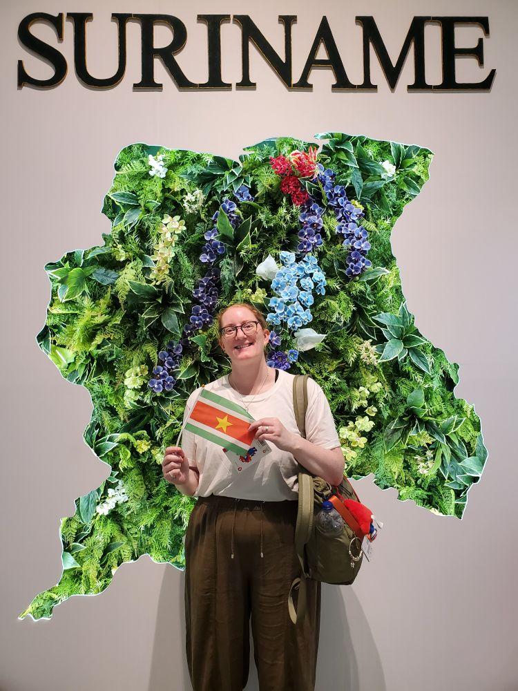 A woman stands in front of a flower wall holding a Suriname flag. The flag is red, white and green stripes with a yellow star in the middle. 