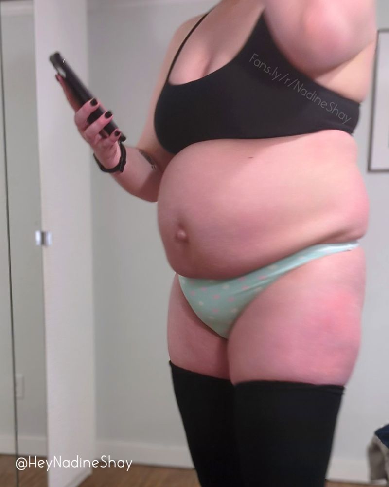 This image is of me, BBW Nadine Shay, a hairy, fat femme standing in profile before a mirror, wearing a black sports bra, a light blue thong, and black thigh-high socks, as I prepare to do a weigh-in video. I'm positioned in front of the viewer, facing the viewer's left, visible from my shoulders to my thighs; my left hand is holding a phone to record the video; my fat, round belly protrudes well beyond my titties and hangs over the waistband of my panties; dimples are visible on my thighs.