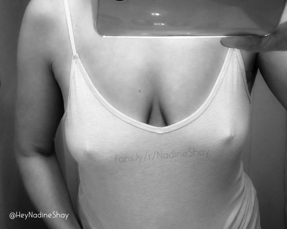 This black and white image is of me - BBW Nadine Shay - a hairy, fat femme wearing a camisole and taking a mirror selfie. I'm positioned in front of the viewer, visible from my shoulders to my belly; my left hand is by my side, and my right hand is holding a phone near my face; my shirt is low-cut and reveals a lot of cleavage; I am braless, and my nipples are erect and poking out against my shirt.