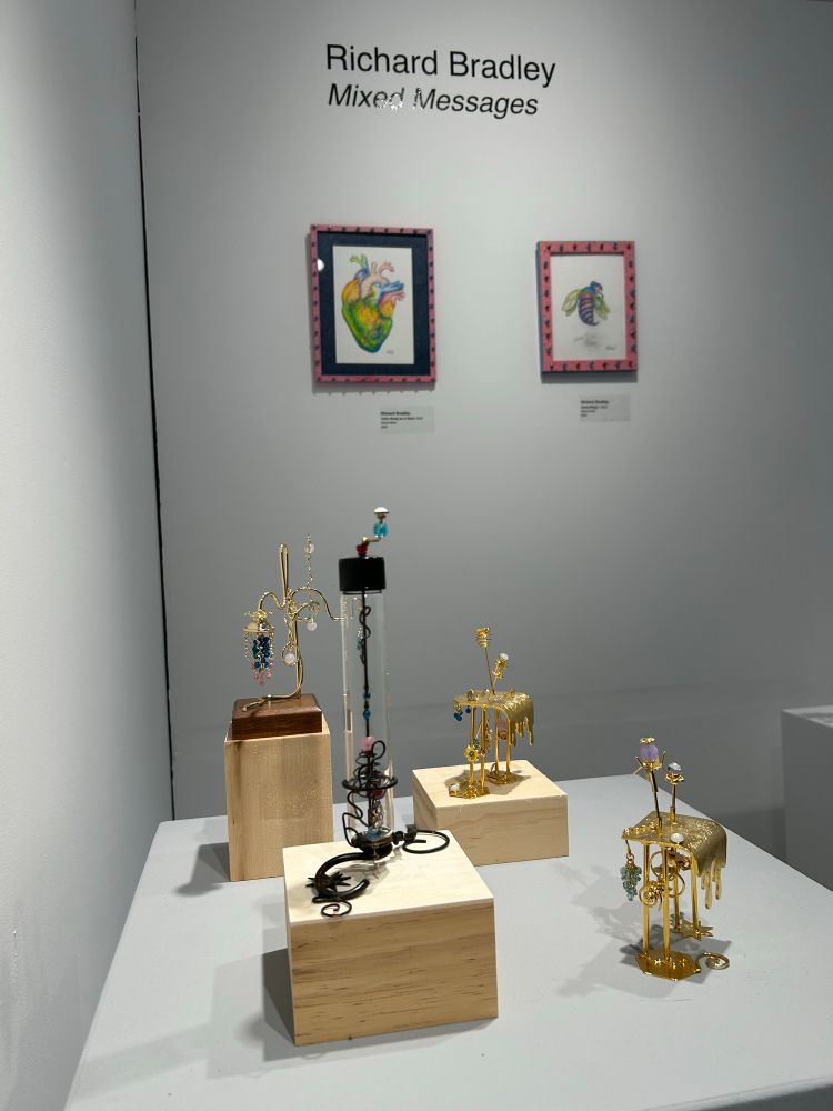 A table top arrangement of four small sculptures by artist Richard Bradley in an art gallery with two of his drawings hanging on the wall behind them