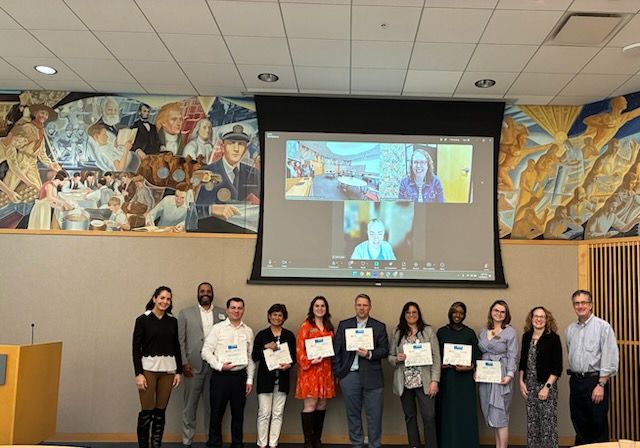 Group of in-person TIARA graduates under a screen that include virtual graduates