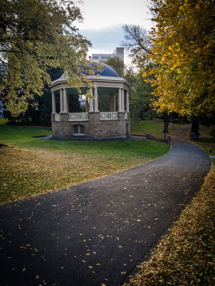 A rotunda sits in a park surrounded by deciduous trees dropping a carpet of brightly coloured leaves. 