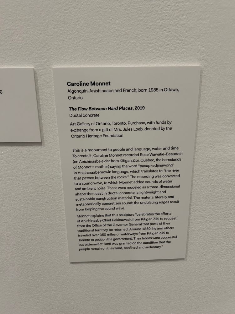 wall text for the sculpture reads:

Caroline Monnet
Algonquin-Anishinaabe and French; born 1985 in Ottawa, Ontario

The Flow Between Hard Places, 2019

Ductal concrete

Art Gallery of Ontario, Toronto. Purchase, with funds by exchange from a gift of Mrs. Jules Loeb, donated by the Ontario Heritage Foundation

This is a monument to people and language, water and time.
To create it, Caroline Monnet recorded Rose Wawatie-Beaudoin (an Anishinaabe elder from Kitigan Zibi, Quebec, the homelands of Monnet's mother) saying the word "pasapkedjinawong" in Anishinaabemowin language, which translates to "the river that passes between the rocks." The recording was converted to a sound wave, to which Monnet added sounds of water and ambient noise. These were modeled as a three-dimensional shape then cast in ductal concrete, a lightweight and sustainable construction material. The material literally and metaphorically concretizes sound: the undulating edges result from looping the sound wave.
Monnet explains that this sculpture "celebrates the efforts of Anishinaabe Chief Pakinawatik from Kitigan Zibi to request from the Office of the Governor General that parts of their traditional territory be returned. Around 1850, he and others traveled over 350 miles of waterways from Kitigan Zibi to Toronto to petition the government. Their labors were successful but bittersweet: land was granted on the condition that the people remain on their land, confined and sedentary."