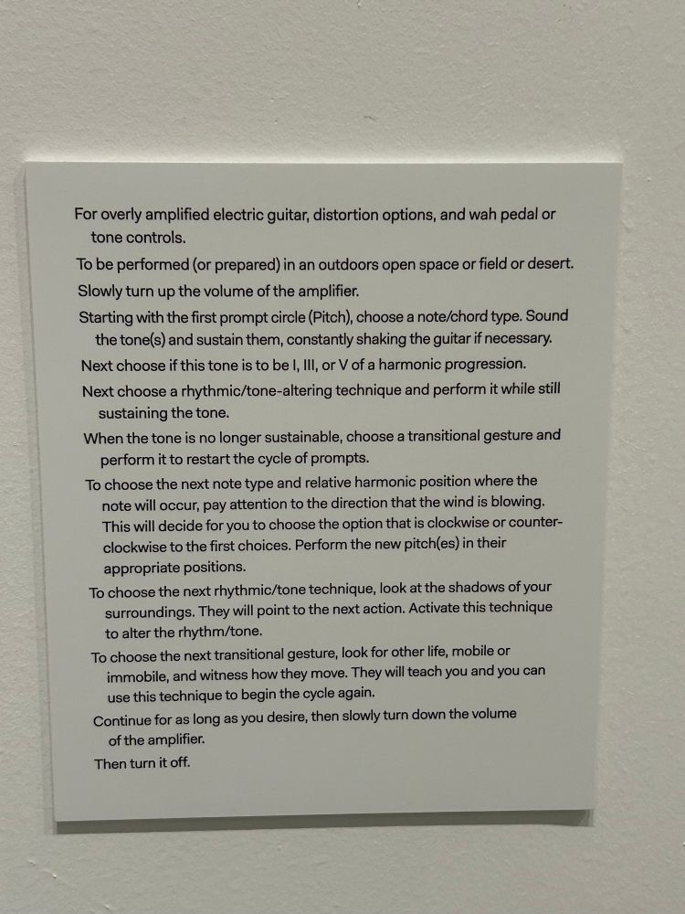 Wall text continues:

For overly amplified electric guitar, distortion options, and wah pedal or tone controls.
To be performed (or prepared) in an outdoors open space or field or desert.
Slowly turn up the volume of the amplifier.
Starting with the first prompt circle (Pitch), choose a note/chord type. Sound the tone(s) and sustain them, constantly shaking the guitar if necessary.
Next choose if this tone is to be I, Ill, or V of a harmonic progression.
Next choose a rhythmic/tone-altering technique and perform it while still sustaining the tone.
When the tone is no longer sustainable, choose a transitional gesture and
perform it to restart the cycle of prompts.
To choose the next note type and relative harmonic position where the note will occur, pay attention to the direction that the wind is blowing.
This will decide for you to choose the option that is clockwise or counterclockwise to the first choices. Perform the new pitch(es) in their appropriate positions.
To choose the next rhythmic/tone technique, look at the shadows of your surroundings. They will point to the next action. Activate this technique to alter the rhythm/tone.
To choose the next transitional gesture, look for other life, mobile or immobile, and witness how they move. They will teach you and you can use this technique to begin the cycle again.
Continue for as long as you desire, then slowly turn down the volume of the amplifier.
Then turn it off.