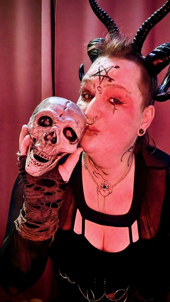 Krissy is a black dress, with demonic horns and a pentacle drawn on her forehead. She is kissing a skull.