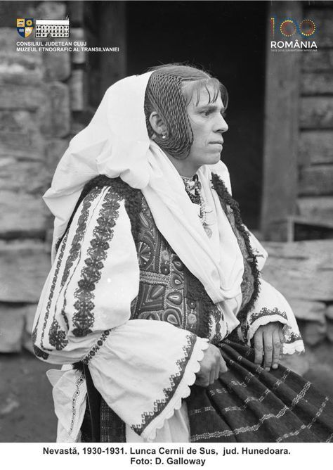 Historical photograph of a Transylvanian peasant woman with white head covering, intricate hair braids, and embroidered vest over a white blouse and skirt. Caption reads, "Nevasta (Wife), 1930-1931. Lunca Cernii de Sus, jud. Hunedura. Foto: D. Galloway."