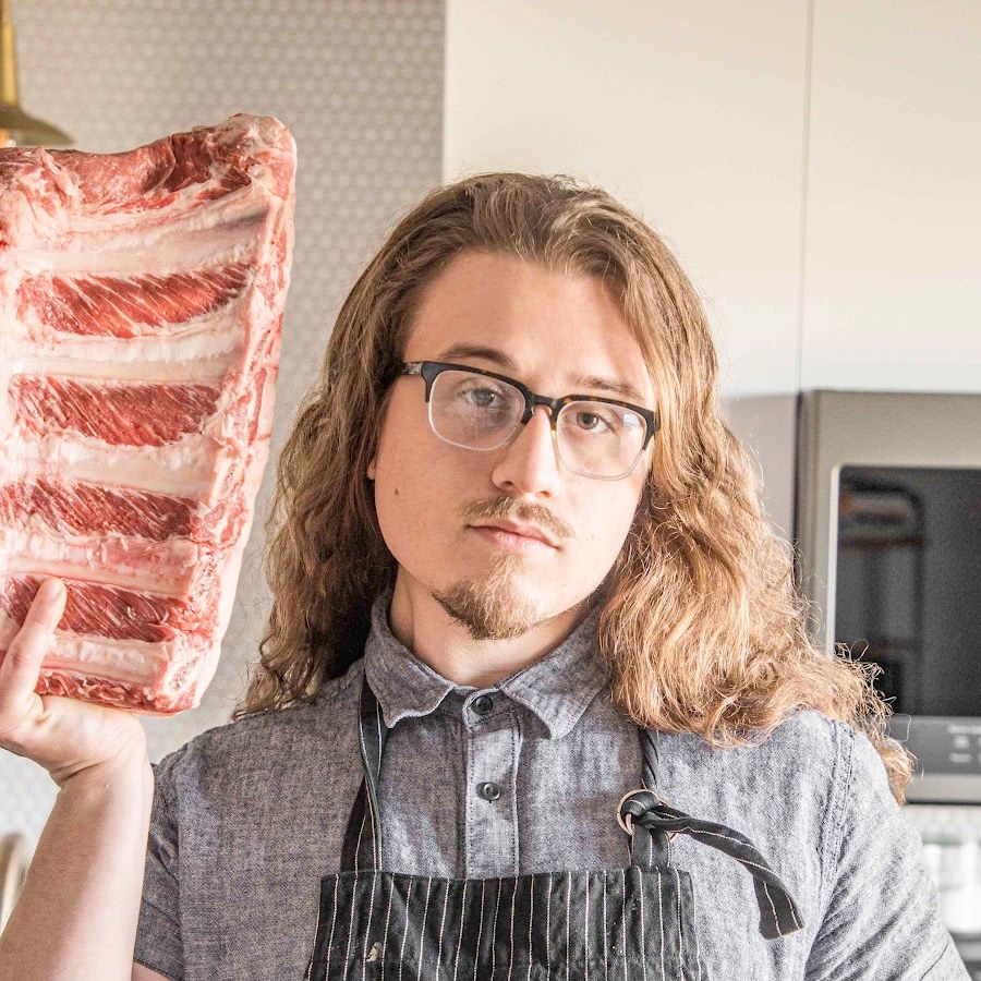 Une photo de Joshua Weissman, avec un tablier. Il porte une énorme poitrine de viande par son bras droit. Il regarde l'objectif, et c’est vrai qu’il ressemble très fortement à Kocobé, avec ses cheveux longs, ses lunettes et sa petite barbichette.