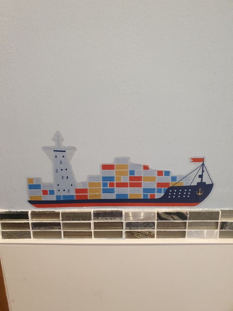 A vinyl sticker of a container ship against a blue-grey wall, sailing on top of a strip of black and silver mosaic tiles above large off-white tiles