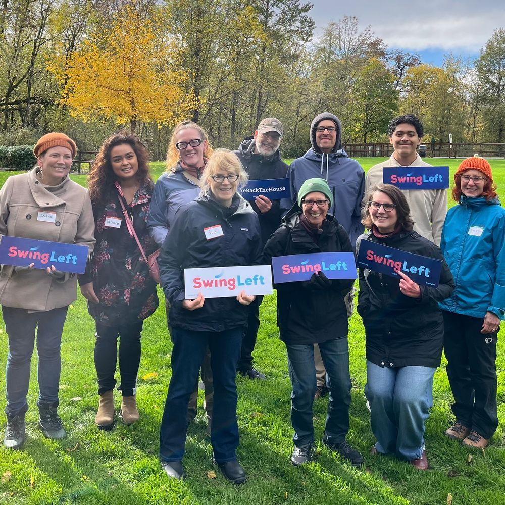 Group of people standing outdoors, smiling, holding "Swing Left" signs on a cloudy day with trees and grass in the background.