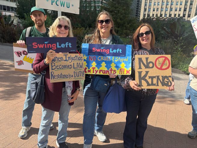 Four people at a protest holding signs with the messages "Swing Left," "When Injustice Becomes Law, Resistance is DUTY," "NO CRUEL & Stupid Tyrants!!!" and "No Kings." They are outdoors on a sunny day in a city setting.
