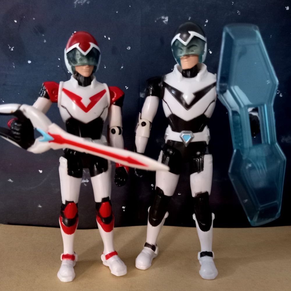 Two official toys of Keith and Shiro in their paladin armor and the same sky background. Keith has his bayard sword and Shiro has a shield. They have their helmets on, but these dolls are known for their wonky faces.