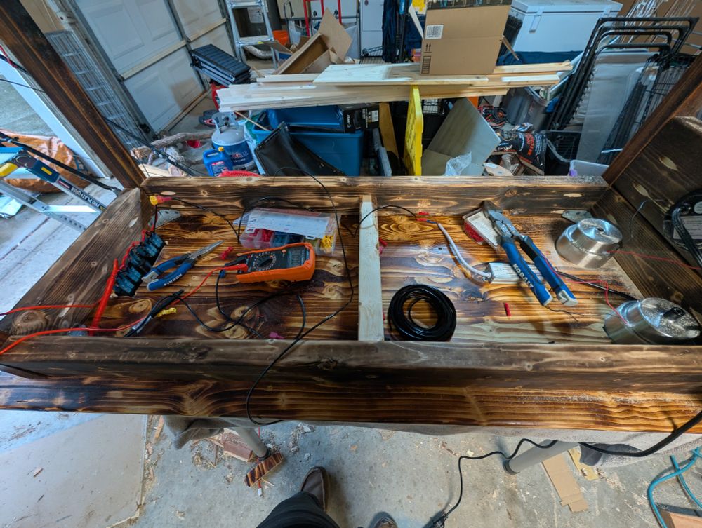 Photo of underside of a dark burned wood coffee table. With wire cutters, lots of cut wire, plyers, and volt meter haphazardly laying about.