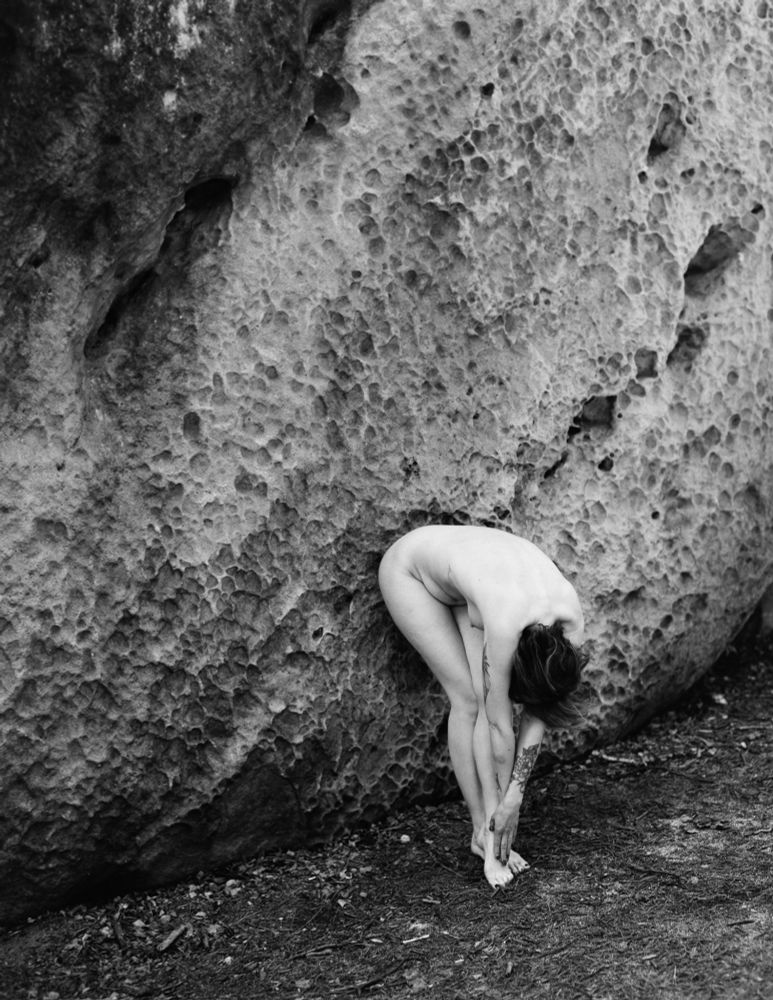 The image depicts a person who is nude and positioned in front of a large, textured rock. They are bending forward, with their arms wrapped around their legs, and their hair falls softly over their head. The contrast between the human figure and the rugged surface of the rock emphasizes themes of vulnerability and connection to nature. The black-and-white photography enhances the mood, lending a timeless quality to the scene while focusing on the shapes and lines formed by both the body and the rock. The overall atmosphere is one of introspection and natural beauty.
