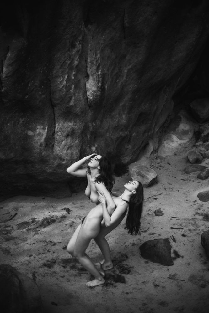 The image features two figures in a close embrace, posed against a backdrop of large, rugged rocks by a sandy area. Both individuals are nude, with one figure standing and gently supporting the other, who appears to be leaning back slightly, creating a dynamic interaction between them. Their hair flows around them, adding movement and softness to the composition. The contrast between the smooth skin of the figures and the rough texture of the rocks enhances the visual interest. The black and white treatment emphasizes shadows and highlights, creating a dramatic atmosphere that underscores their connection. The overall mood conveys intimacy, tension, and a sense of unity against a powerful natural backdrop.
