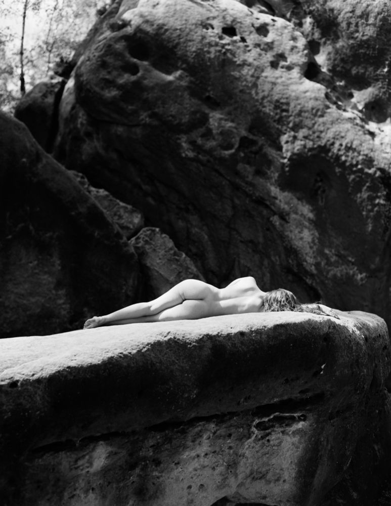 This arresting black and white photograph presents a scene of striking contrasts. The central focus is a nude woman lying prone on a large, weathered rock ledge. Her body curves gently against the rough texture of the stone, conveying a sense of both vulnerability. Her posture is relaxed, with one arm extended along her body, creating a line of graceful contrast against the starkness of the rock. The setting is deeply integrated with the subject. The woman rests on a prominent rock formation within what appears to be a heavily wooded area. Behind her rises a sheer rock face. The natural setting dwarfs her form, emphasizing the power and permanence of the environment. The photograph is rendered in stark black and white, which strips away any distraction of color and amplifies the texture and tonal range of the scene. The lighting seems relatively even, highlighting the details of the woman’s body and the patterns etched into the rock. This deliberate aesthetic choice lends a timeless, almost surreal
quality to the image, elevating it beyond a simple portrait and inviting contemplation on themes of nature, human connection, and the passage of time.