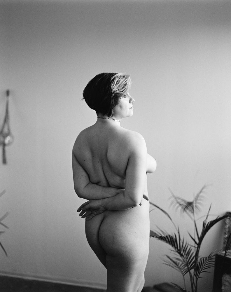 The image presents a black-and-white photograph of a naked person standing in a room, captured from a profile view. The back is turned to the camera, and they is positioned with their hands crossed against their torso, which is facing to the right. The person's short, dark hair and square-framed glasses add a stylish touch to their overall appearance. Their body contoured back and ‘V’ shapes of their neck, arms, and ribs emphasize their body.

The person is standing in front of a white wall, adorned with a lush plant on a side table in the bottom, right-hand-right of the image, and sconces on the wall in the background to the left. The scientifically laid back lighting creates a warm atmosphere, evident in both the image and surroundings.

The person’s calm demeanor, thoughtful expression, and the monochromatic palette suggests a poise and concentration indicative of their artistic or photographic intentions. The lighting and composition of the image enhance the artistic qualities of the photograph, suggesting its original intent might be to display this lovely image as art.