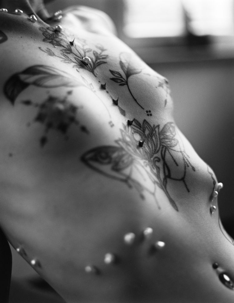The photograph is a close-up, black and white portrait focusing on the upper torso and shoulder of a person. The subject appears to be nude, with their body angled slightly to reveal a side view. The skin tone is soft and appears smooth, accentuated by the diffused lighting.
The most striking feature is the intricate and expansive floral tattoo that covers a large portion of the upper back and shoulder. The tattoo design is detailed, incorporating a variety of floral elements and delicate line work. It has a somewhat ethereal, flowing quality.
Adding a unique and striking element, small pearls are adhered to the skin, creating a delicate and subtly
shimmering effect. These are sparsely distributed across the shoulder and upper back, drawing attention to their placement and creating a textural contrast against the smooth skin and detailed tattoo.
The background is softly blurred, suggesting a window or wall with diffused light filtering through. This creates a gentle, almost dreamlike atmosphere. The overall mood is intimate, emphasizing the beauty and detail of the body art, the skin, and the unexpected addition of the pearls. The composition draws the eye to the interplay of light, shadow, and the delicate intricacy of the tattoo design, now accented by the subtle shimmer of the pearls.