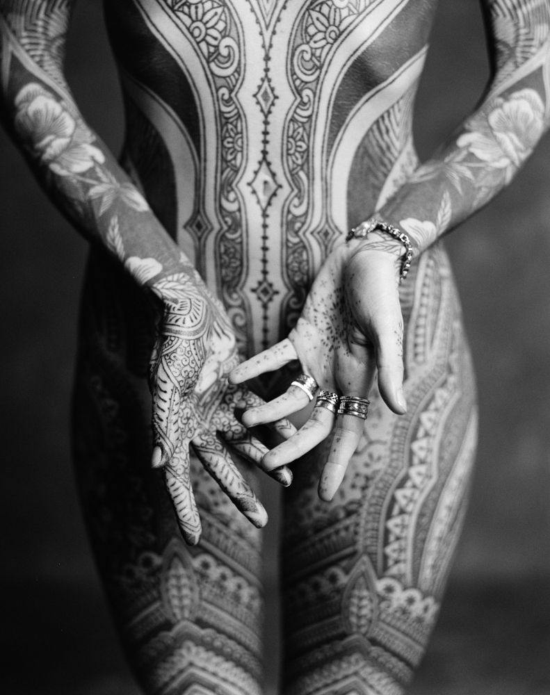 The photograph presents a striking, close-up view of a person's chest and torso. The composition focuses on the front of the body, revealing a vast expanse covered in intricate and detailed tattoos. The design incorporates a complex blend of floral and geometric patterns, showcasing a unified and continuous artwork across the body.

The skin itself appears smooth and taut, serving as a canvas for the meticulously applied ink. The tattoos are dense and intricate, demonstrating a significant commitment to the body modification.

The person's hands are clasped gently in front, highlighting the rings adorning their fingers. The fingers themselves are prominently featured, revealing the detail of their texture and the placement of the jewelry. The lighting is carefully controlled, creating subtle shadows and highlights that accentuate the contours of the body
and the depth of the tattoo designs. The backdrop is dark, ensuring the focus remains entirely on the artwork and the individual's form. The overall impression is one of artistic intention and a deliberate focus on the body as a medium for art.