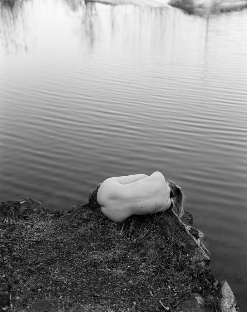 The photograph presents a single figure reclining on a dark, muddy bank overlooking a body of water. The figure, appearing nude, is positioned horizontally, with their face turned away from the viewer, gazing out towards the expanse of water. Their hair is visible, cascading around their head and shoulders.
The bank itself is composed of dark, damp earth and scattered vegetation, creating a stark contrast to the lighter tone of the figure’s skin. The water fills the majority of the frame, its surface rippled with gentle waves that create a sense of movement and reflection. The reflection of the figure can be faintly observed on the water’s surface, blurring the boundary between reality and illusion.

The overall composition is minimalistic and serene, evoking a feeling of solitude and contemplation. The
monochrome palette emphasizes the textures and forms, contributing to a quiet, dreamlike atmosphere. The photograph’s simplicity draws attention to the relationship between the individual, the natural environment, and the illusion of reflection.