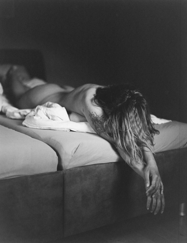 The image portrays a serene scene in which a person is lying face down on a bed. Their body is partially covered by a bedspread, and their hair cascades down, creating a sense of intimacy and vulnerability. The lighting appears soft and gentle, enhancing the mood of tranquility. The focus seems to be on the relaxed posture and the sense of peace in the space.
