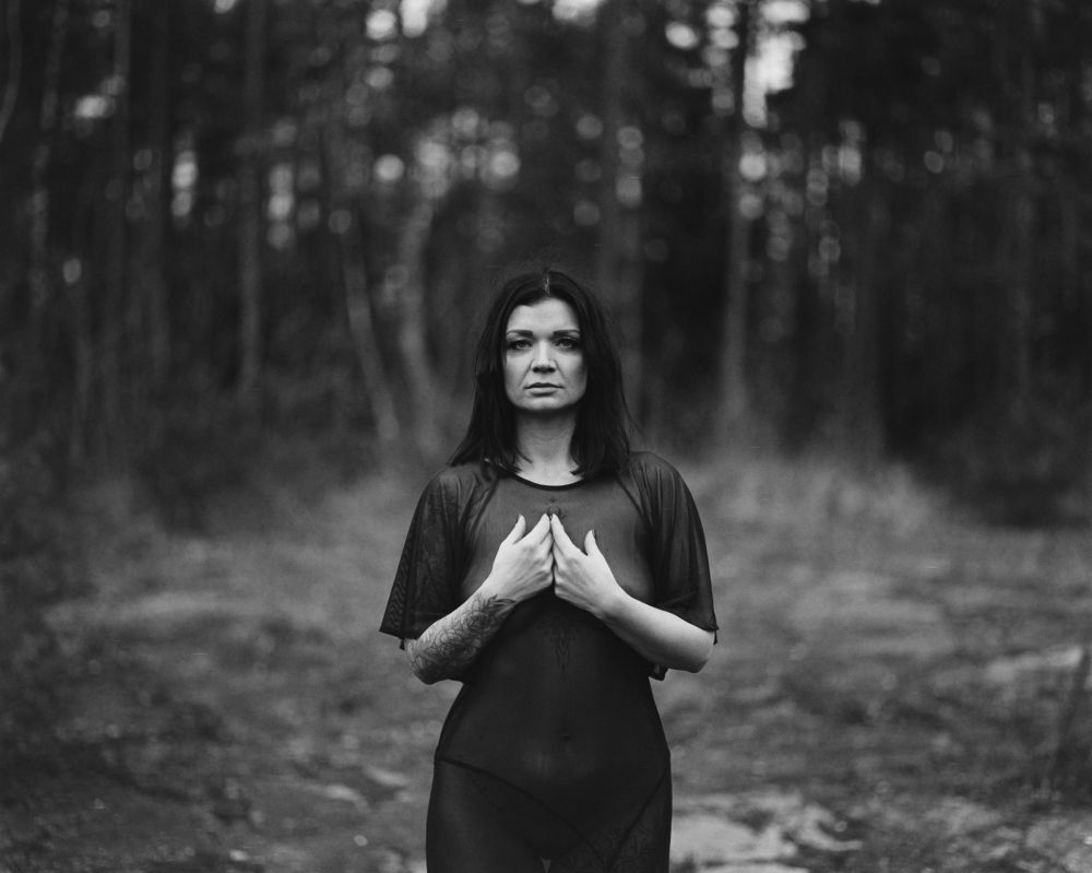 The image features a person standing in a forested area, looking directly at the camera with a strong and serious expression. They're wearing a sheer black top that subtly highlights their figure and tattoos, conveying a sense of strength and vulnerability simultaneously. The background is blurred, with tree trunks and foliage blending into an indistinct, shadowy backdrop, adding depth to the scene. The black-and-white style enhances the emotional intensity of the portrait, focusing on the subject's gaze and posture, which suggests both confidence and introspection.
