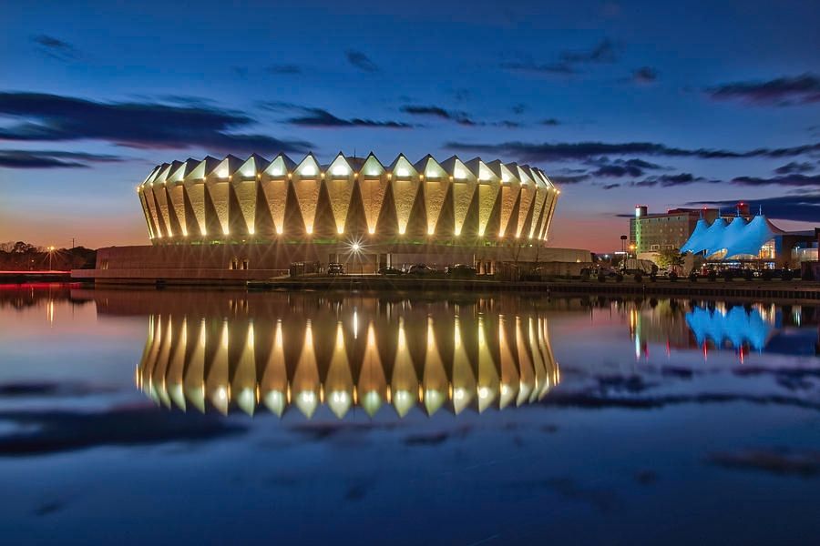 An image of Hampton Coliseum at sunset reflected in the water next to it; it is lit up so it looks even more like a UFO than during the day 