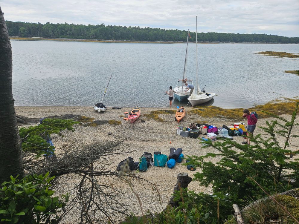 Two small sailboats, 3 kayaks, and camping gear on a small beach in Casco Bay, Maine.